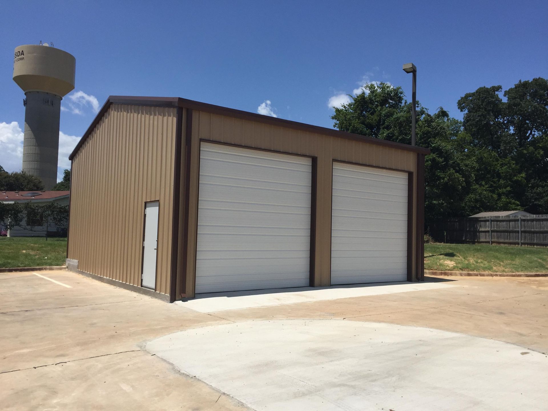 Tan metal garage with two white roll-up doors and a side access door, near a water tower under a blue sky.