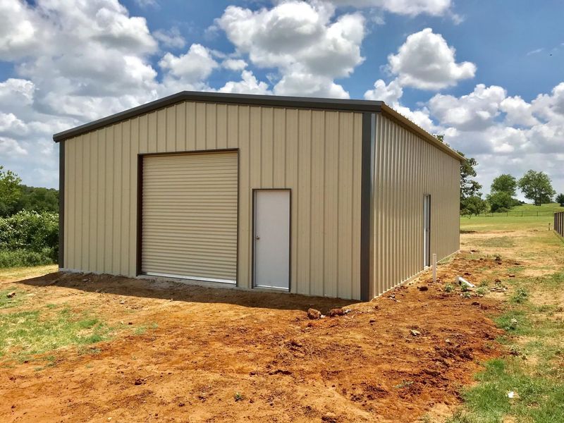 A beige metal warehouse with a roll-up door and a side entry door, set on a dirt lot under a blue sky with clouds.