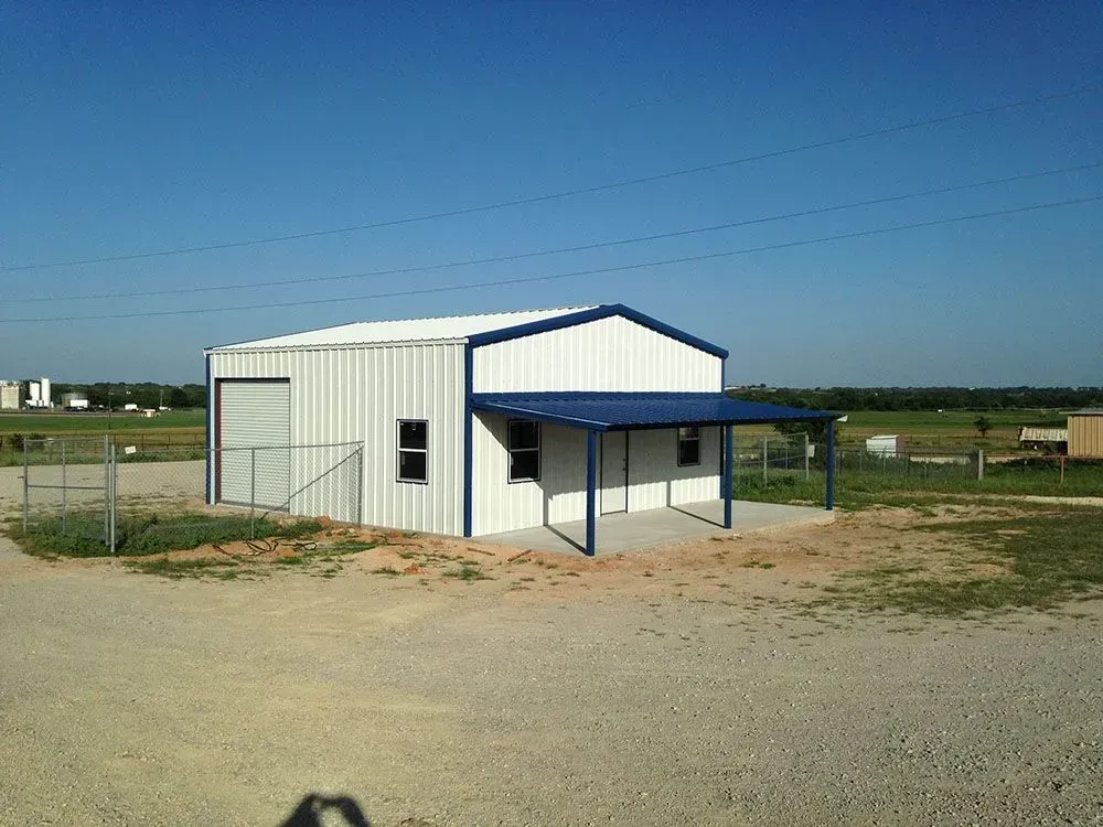 A white metal building with blue trim, a large garage door, and a covered porch on a gravel lot under a clear blue sky.
