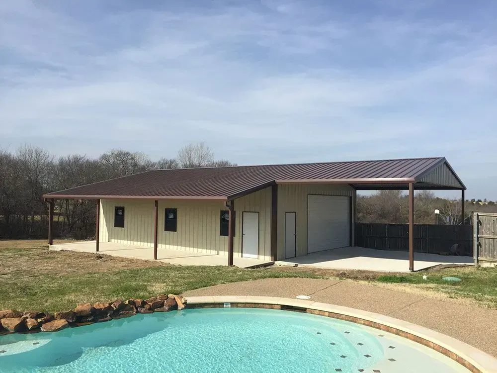 A metal-sided workshop building with a porch and carport sits beside a swimming pool on a sunny day.