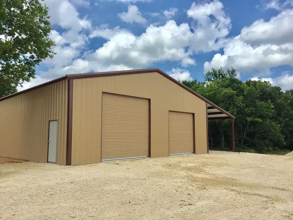 A tan metal building with two large garage doors, a walk-in door, and an open side porch against a cloudy blue sky.