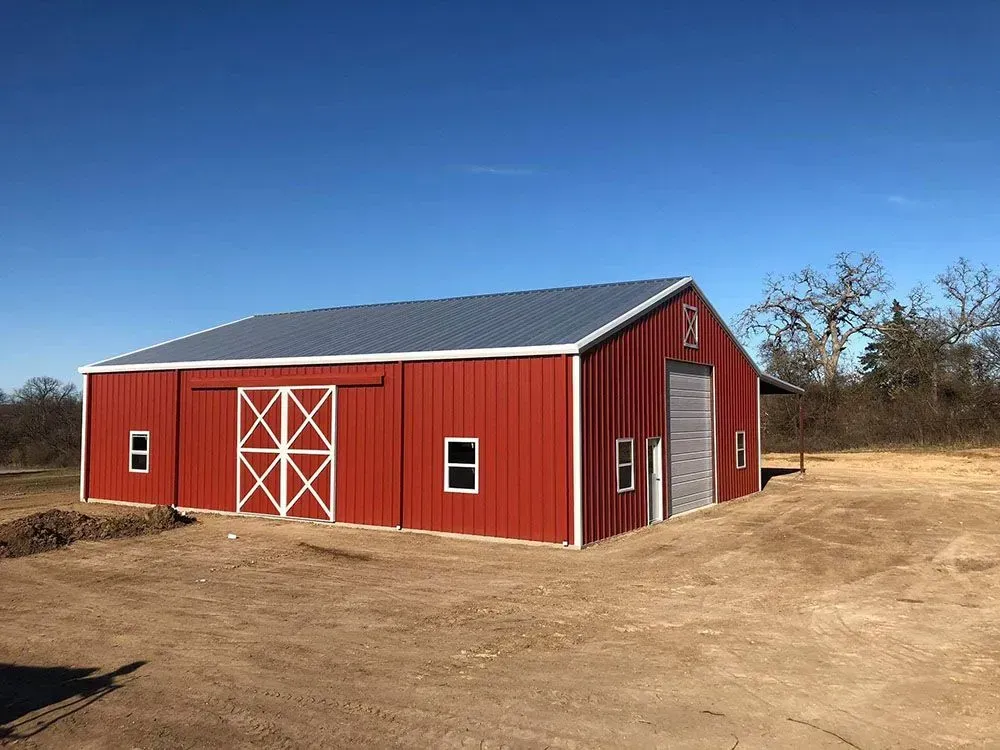 A red metal barn with white trim, sliding doors, and a grey roof sits on a dirt lot under a clear blue sky.