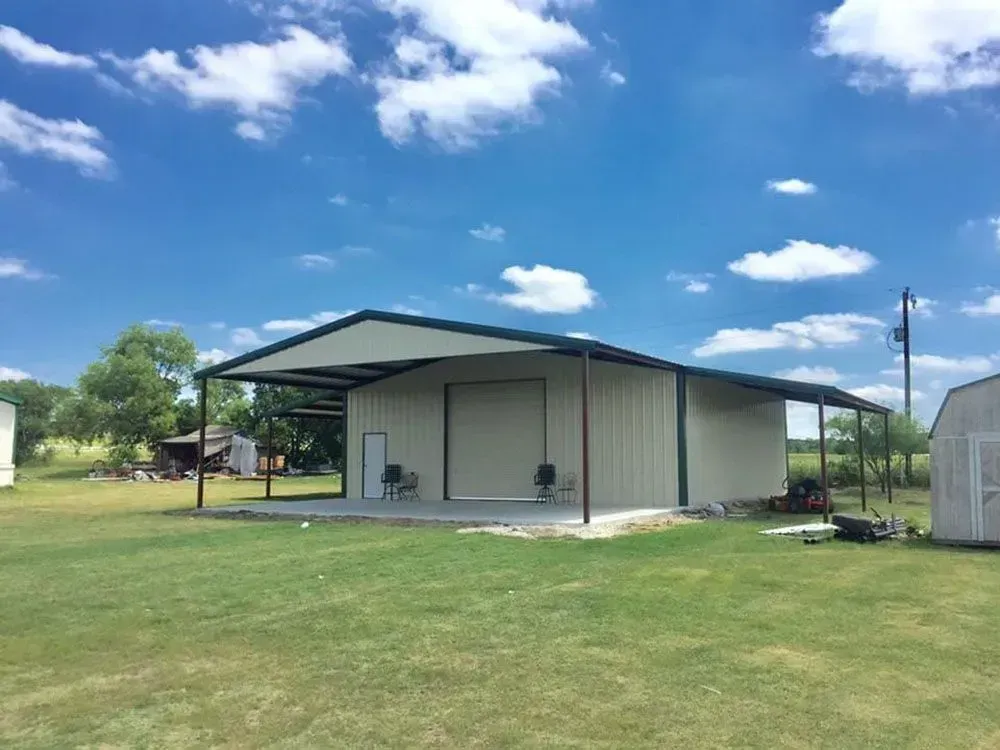 A beige metal building with an attached carport sits on a grassy field under a bright blue, partly cloudy sky.