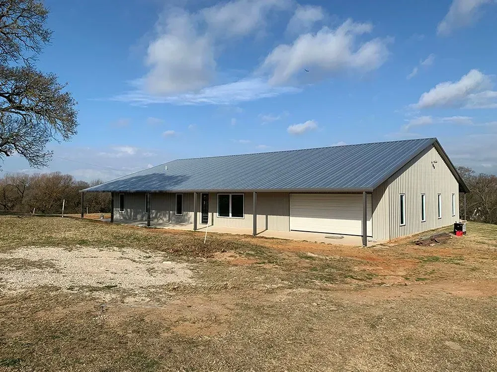 A newly constructed, single-story metal building with a gray roof, tan siding, and a white garage door on a grassy lot.