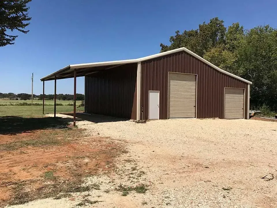 A brown metal workshop with a side lean-to, a roll-up door, and a white walk-in door on a gravel lot under a blue sky.