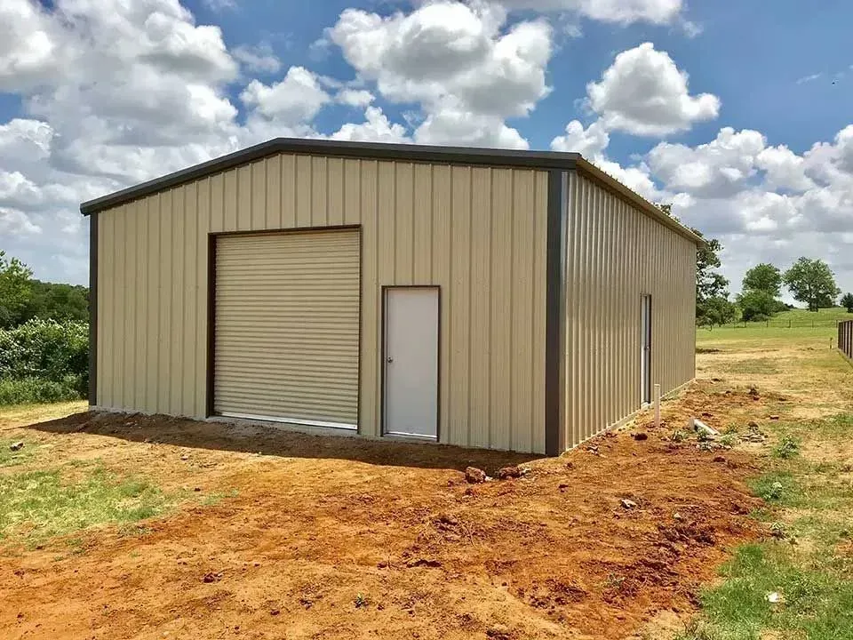 A tan metal workshop with a roll-up door and a single door on a dirt lot under a blue sky with clouds.