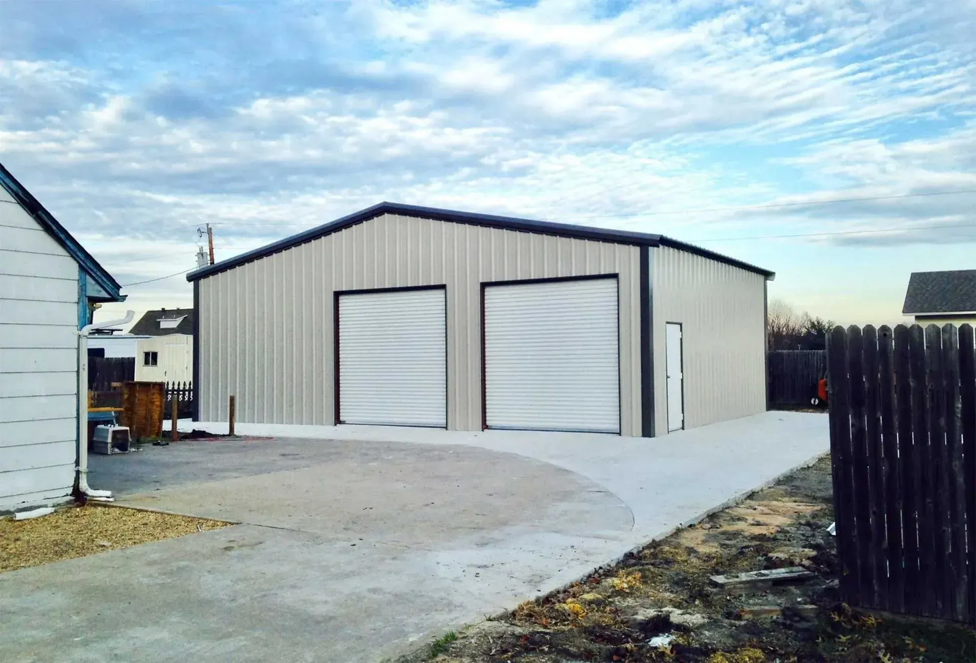 A beige, metal-sided detached garage with two white roll-up doors and a side entry door, set on a concrete driveway.