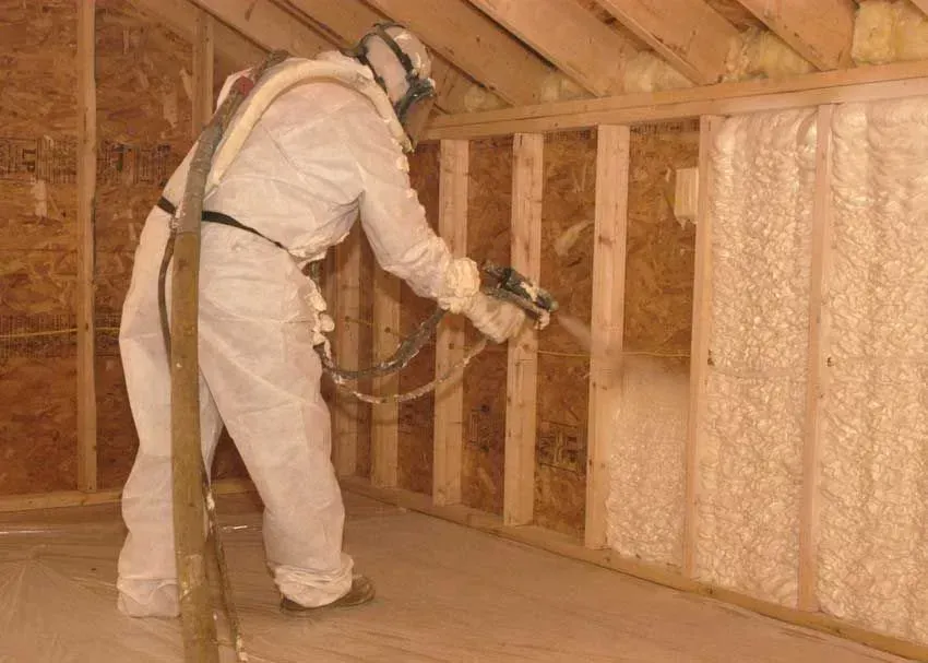 A worker in protective gear sprays foam insulation into the wood-framed wall of an unfinished attic.