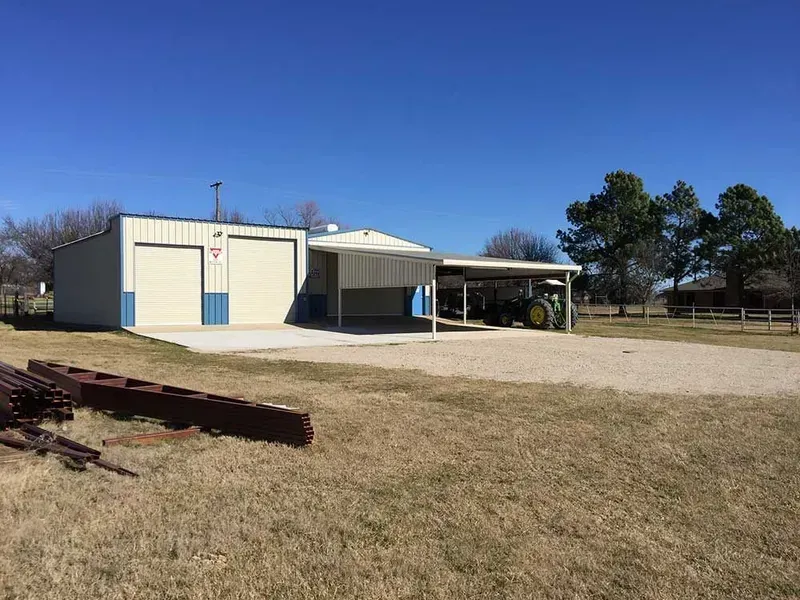 A light-colored metal workshop with two garage doors and a wide, covered equipment shed under a clear blue sky.