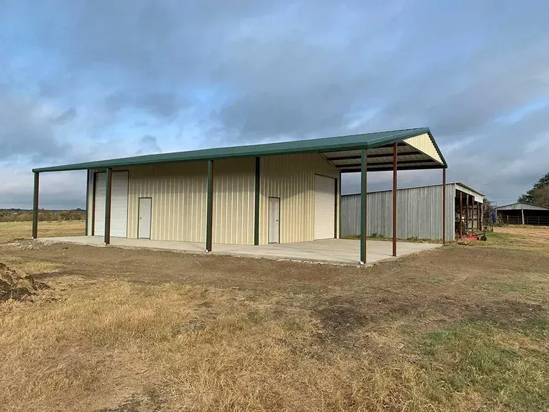 A tan metal building with a dark green roof and an open-sided carport, situated on a dry, grassy rural property.