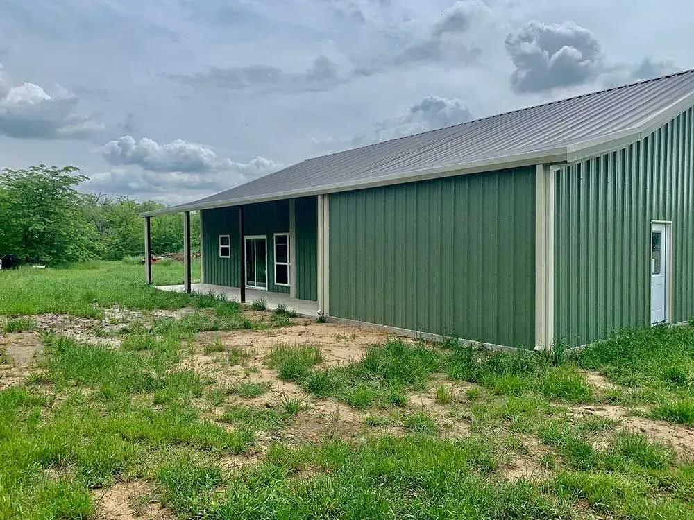 A green metal-sided building with a covered porch and grey roof sits on a grassy lot under a cloudy sky.