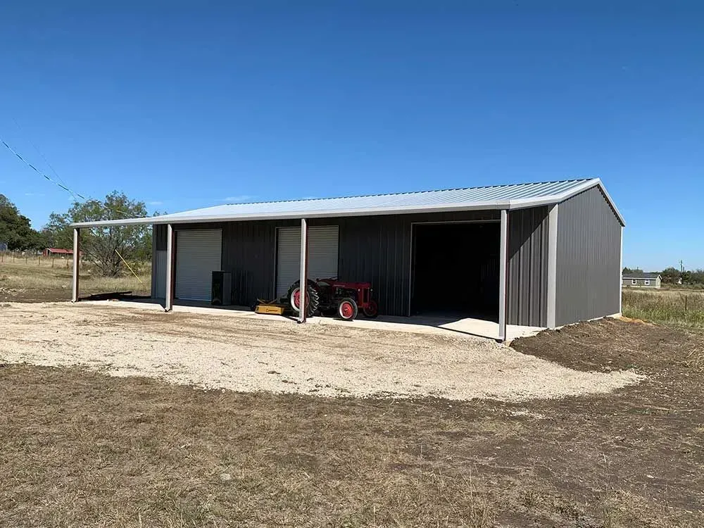 A gray metal barn with an open front, a gravel driveway, and a red tractor parked in front under a clear blue sky.