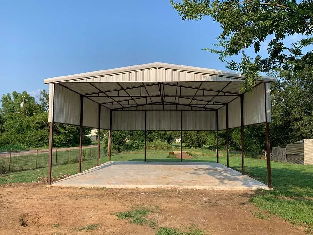 A light-colored metal carport structure with an open front stands on a concrete pad in a grassy, outdoor area.