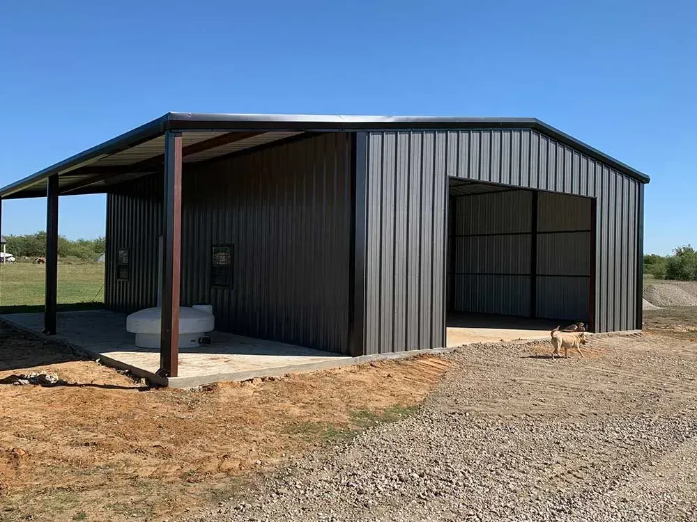 A dark metal barn with an open garage bay and a covered side patio sits on a gravel lot under a clear blue sky.