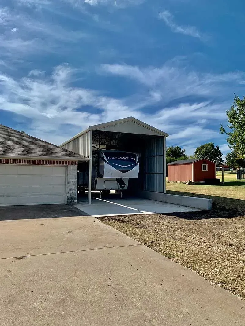 An RV parked under a metal carport next to a house, with a small red shed in the grassy background under a blue sky.