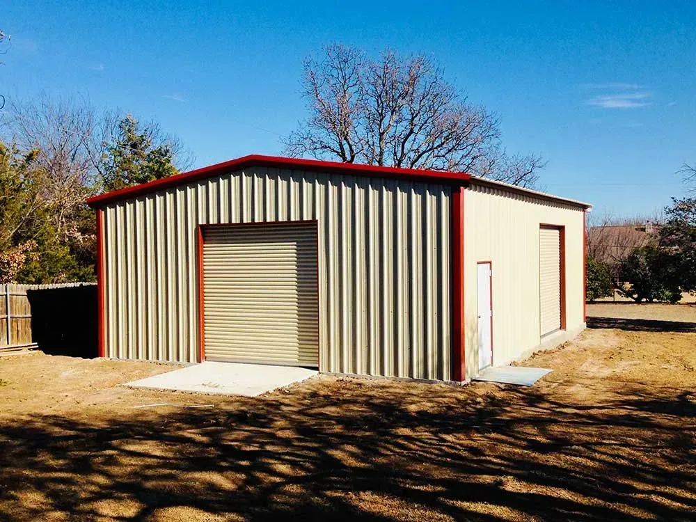 A light-colored metal storage building with red trim, a roll-up door, and a single pedestrian door in a grassy lot.