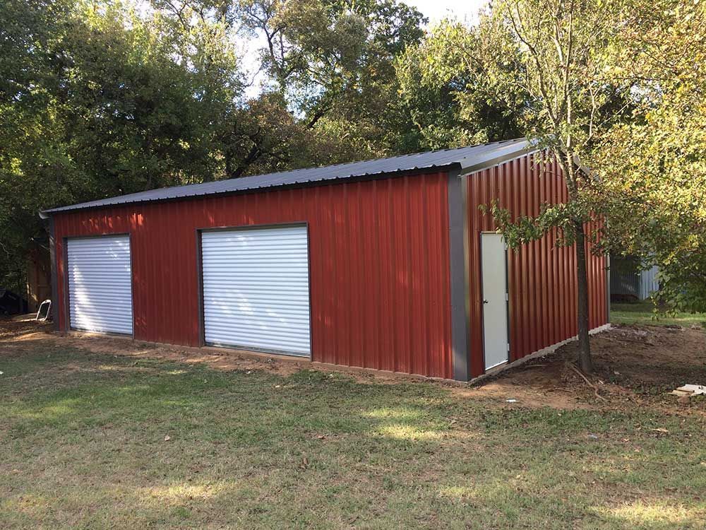 A red metal workshop with two white roll-up doors and a side entry door, situated on a grassy lot surrounded by trees.