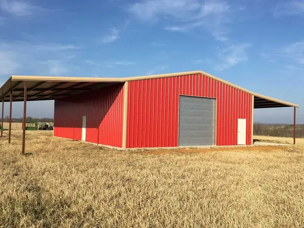 A red metal barn with a tan trim and roof extensions on both sides, situated in a dry, grassy field under a blue sky.