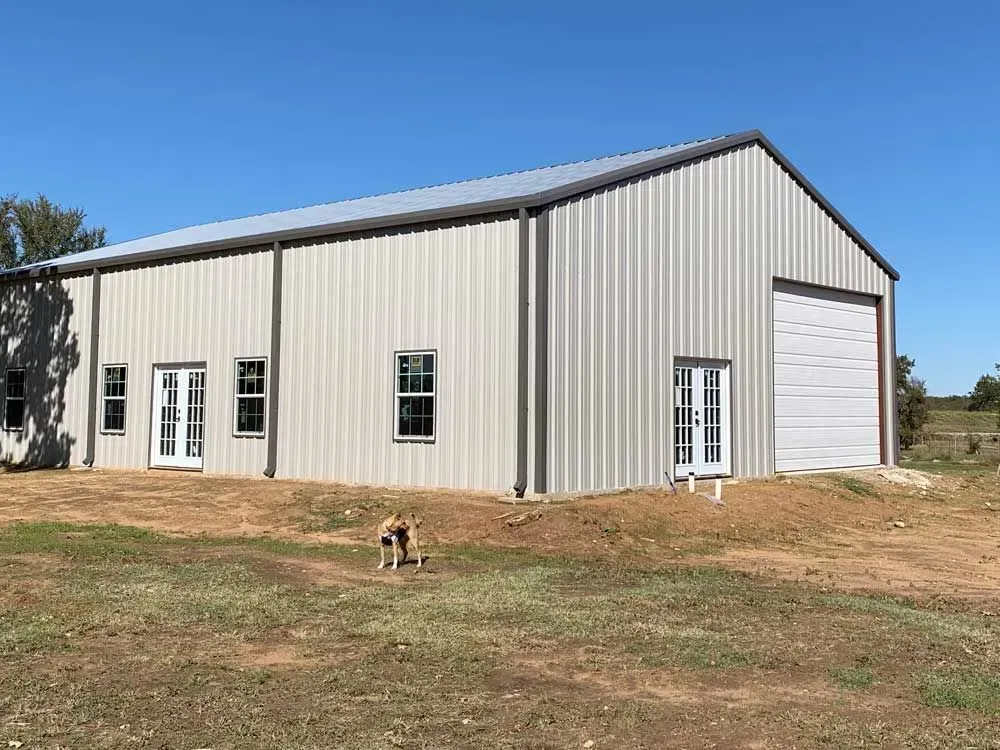 A light-gray metal pole barn with white doors and windows on a grassy lot, with a small dog standing in the foreground.