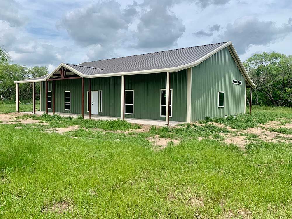 A green metal-sided building with a dark metal roof, a front porch, and several windows, set in a grassy, open field.