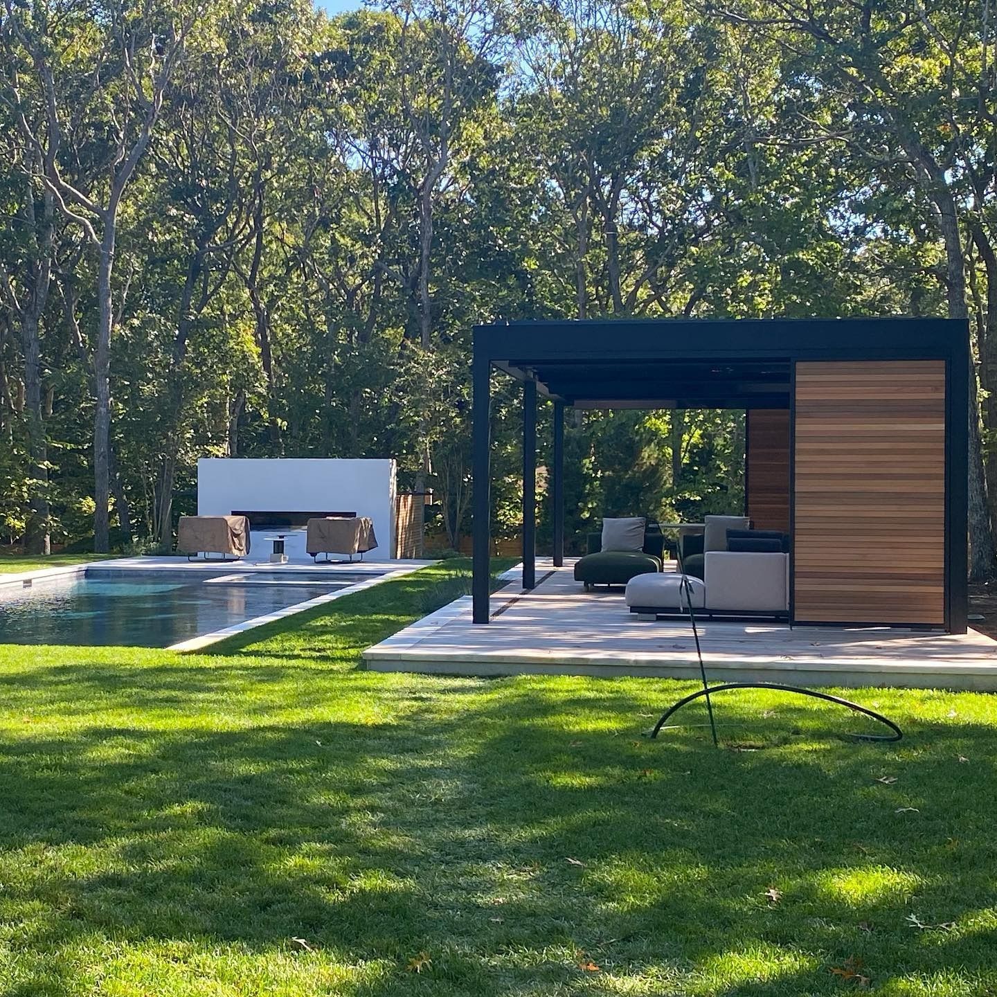 Backyard with pool, patio, and modern structures. Green lawn, trees in background, sunny day.