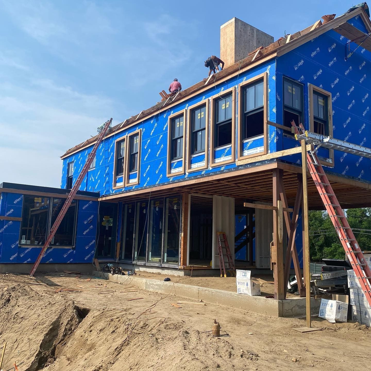 Construction of a two-story house, blue wrap, workers on roof, ladders, clear day.