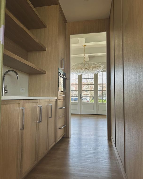 Narrow, light-wood-paneled hallway with cabinetry, shelves, and oven. The hallway leads to a room with French doors and a chandelier.