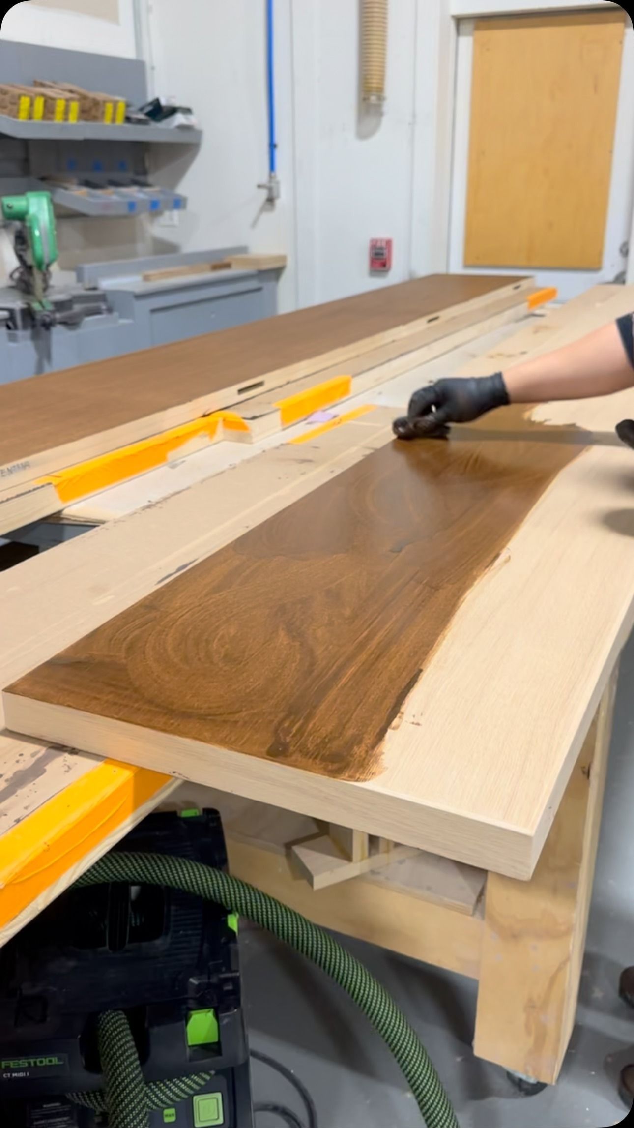 A person wearing gloves applies a dark wood stain to a light-colored wooden slab in a workshop.