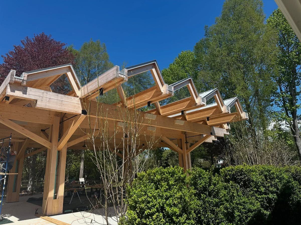 Wooden pergola with zigzag roof, glass panels, greenery, and blue sky.