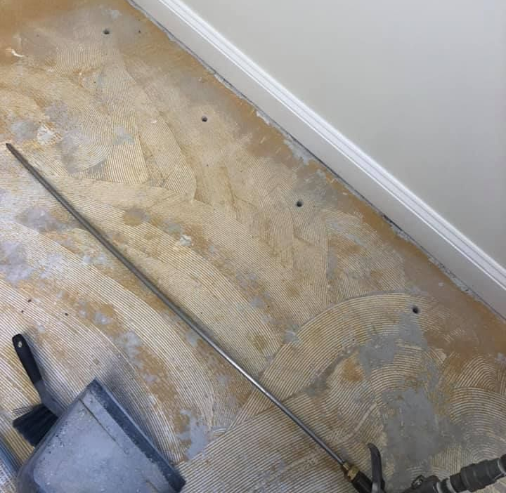 Floor with adhesive residue and several holes, a metal tube, and a bucket. White wall and molding.