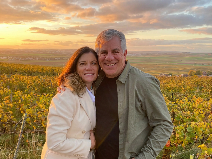 Couple smiling in vineyard at sunset. Woman in cream jacket, man in green shirt.