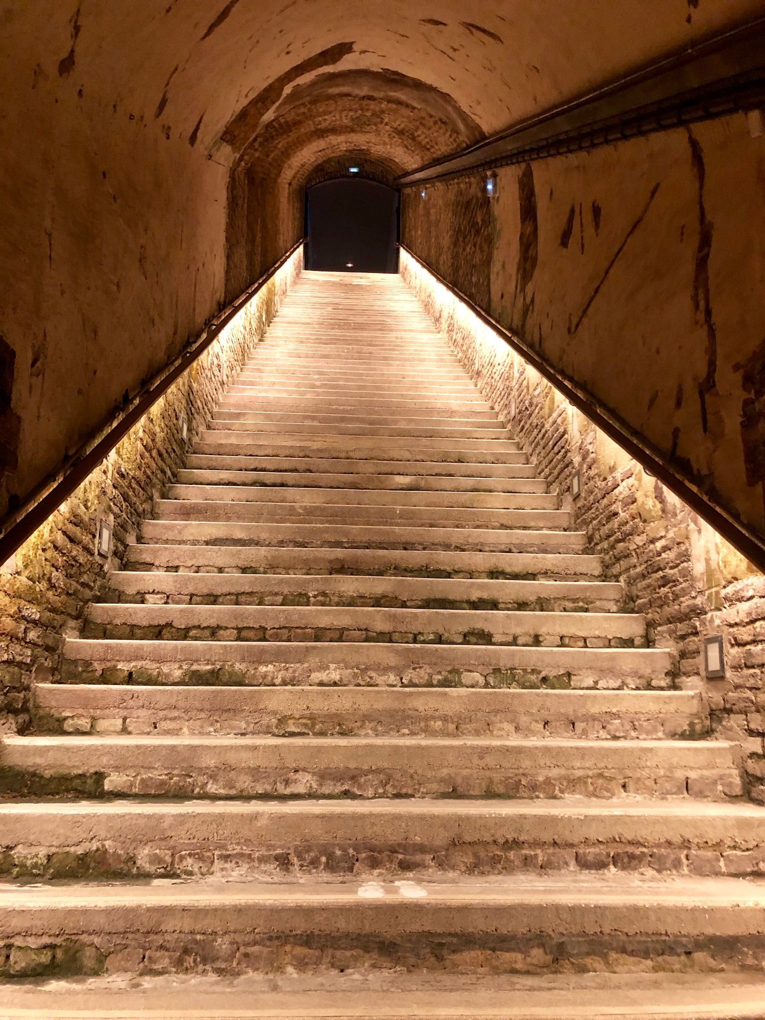 Stone staircase ascending into a dimly lit brick tunnel. Light strips illuminate the stairs and walls.