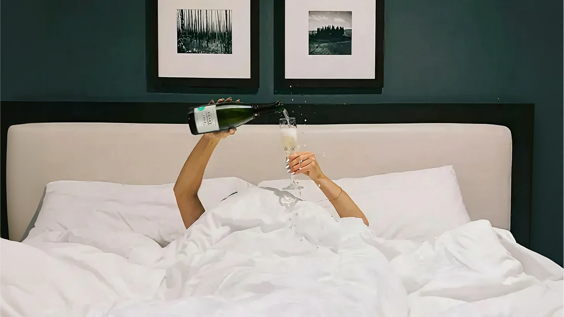 Person pouring champagne into a glass while lying in bed, white sheets, dark headboard, teal wall, framed art.
