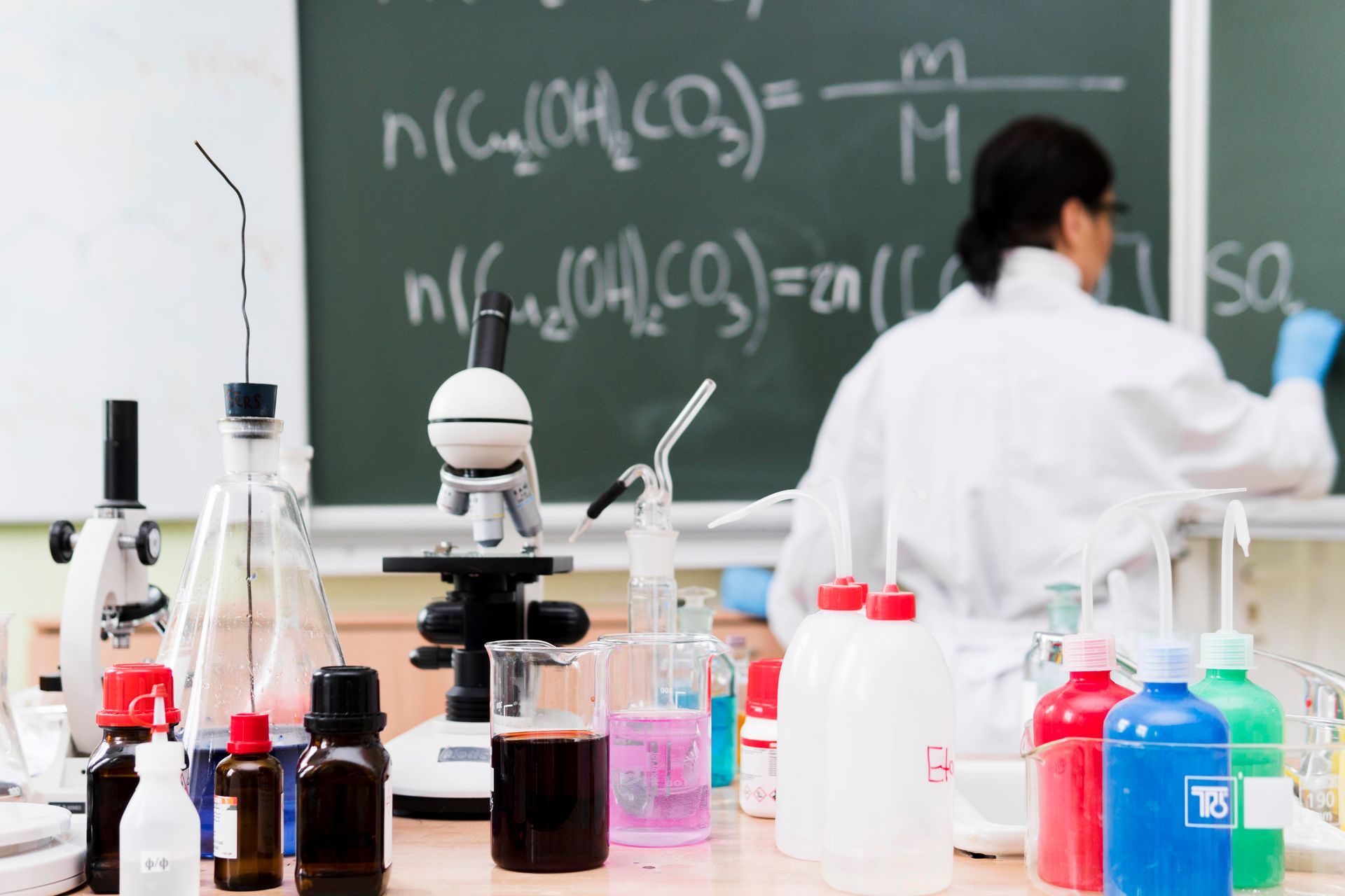 A scientist in a white lab coat writes chemistry formulas on a chalkboard behind a lab bench filled with equipment.