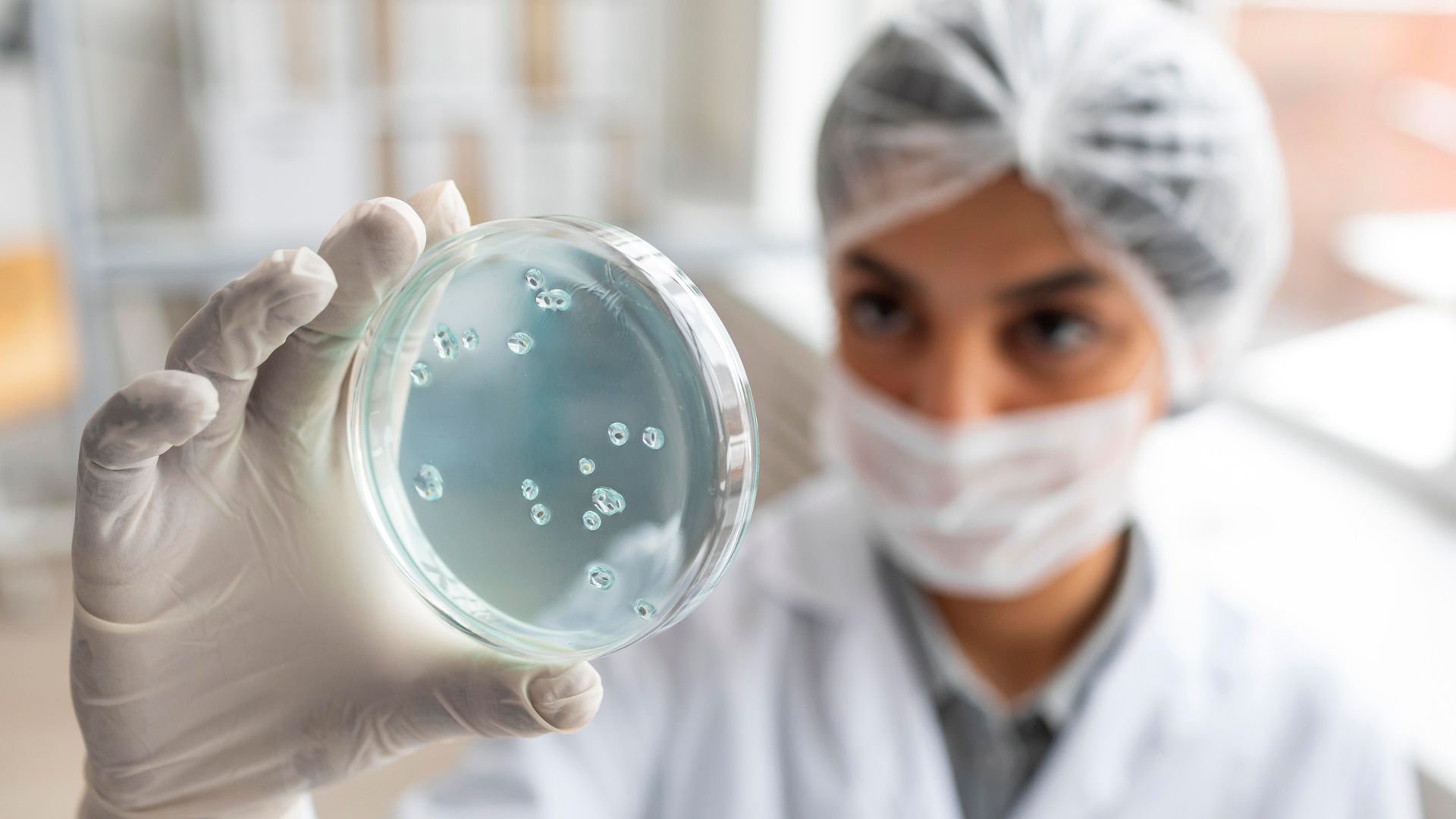 A scientist in a lab coat, mask, and hairnet holds a petri dish containing a bacterial culture for analysis.