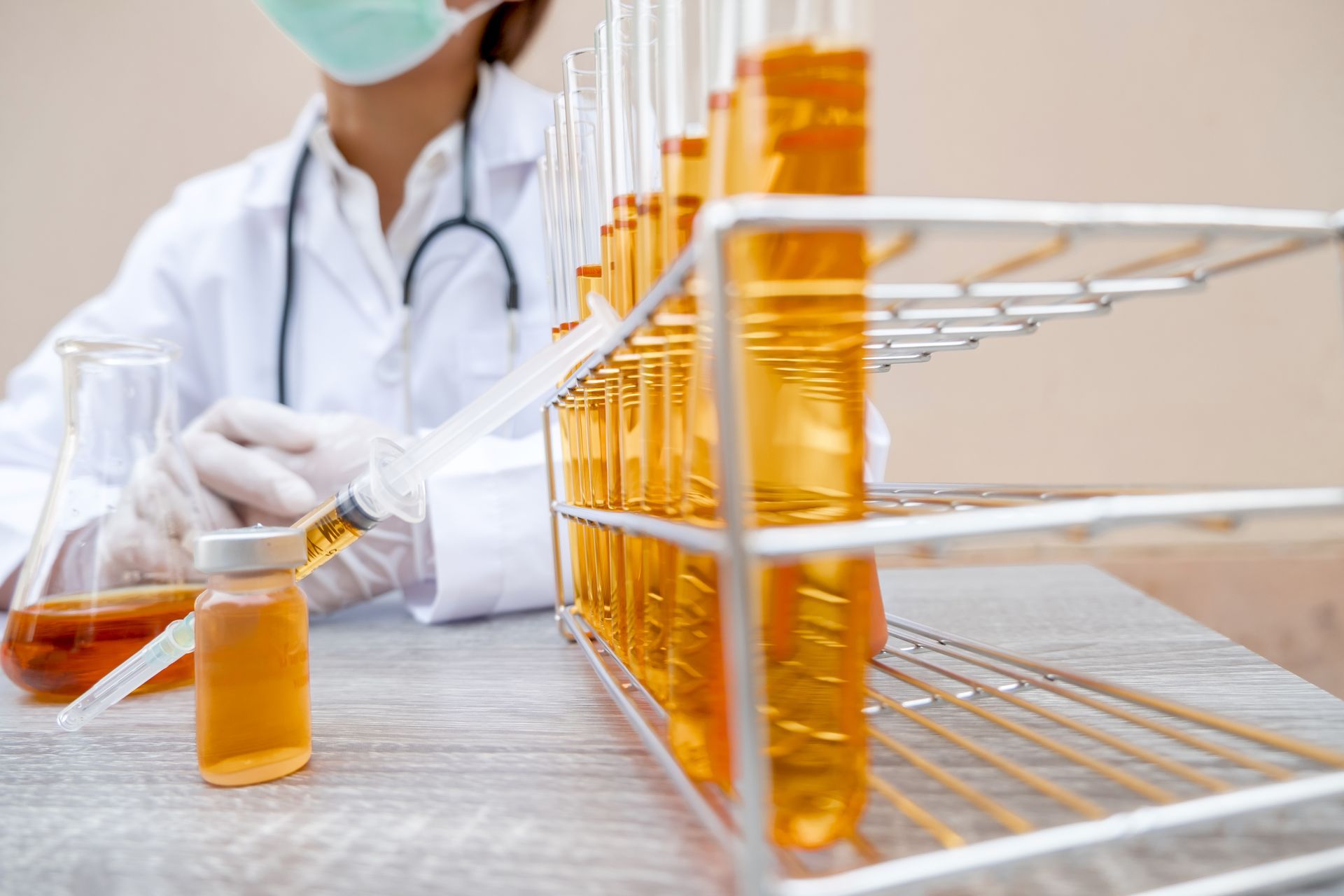 A professional in a lab coat and mask handles a syringe and test tubes filled with a golden-yellow liquid.