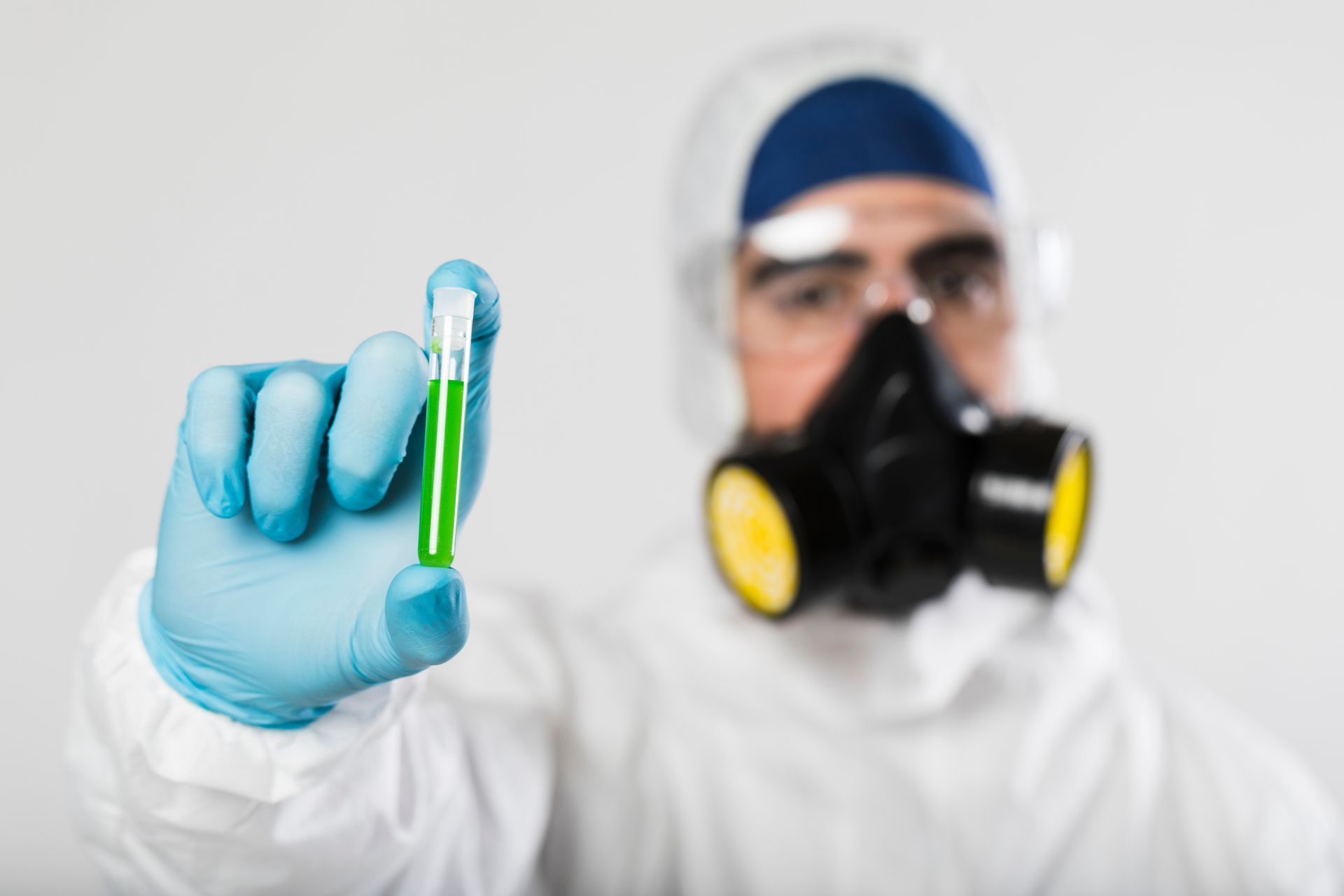 A person in a hazmat suit, mask, and blue gloves holds a test tube filled with green liquid against a white background.