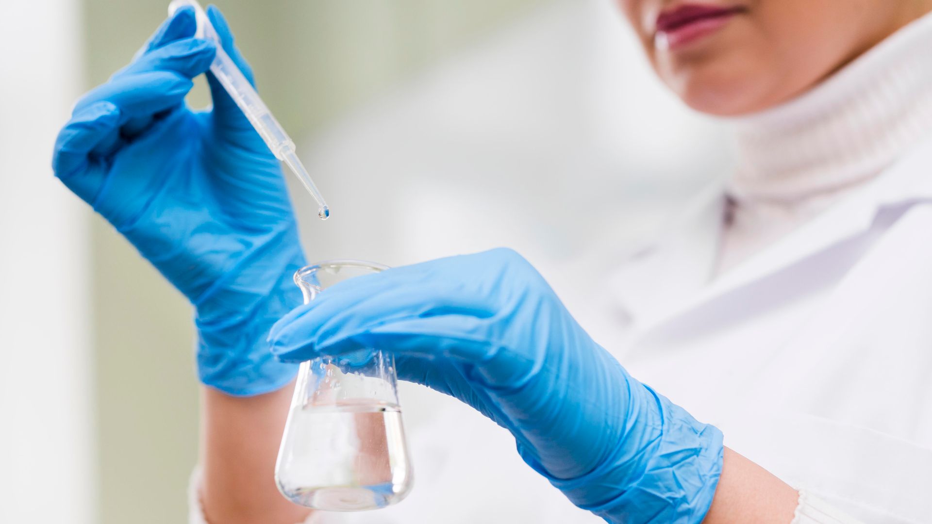A person wearing a lab coat and blue gloves uses a dropper to add liquid to a glass flask in a laboratory setting.