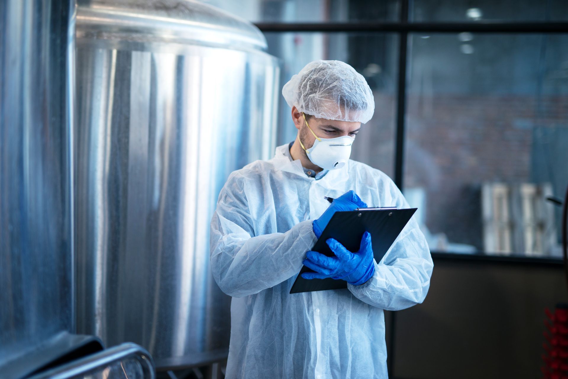 A worker in protective clothing, hairnet, and mask takes notes on a clipboard in front of large metal industrial tanks.