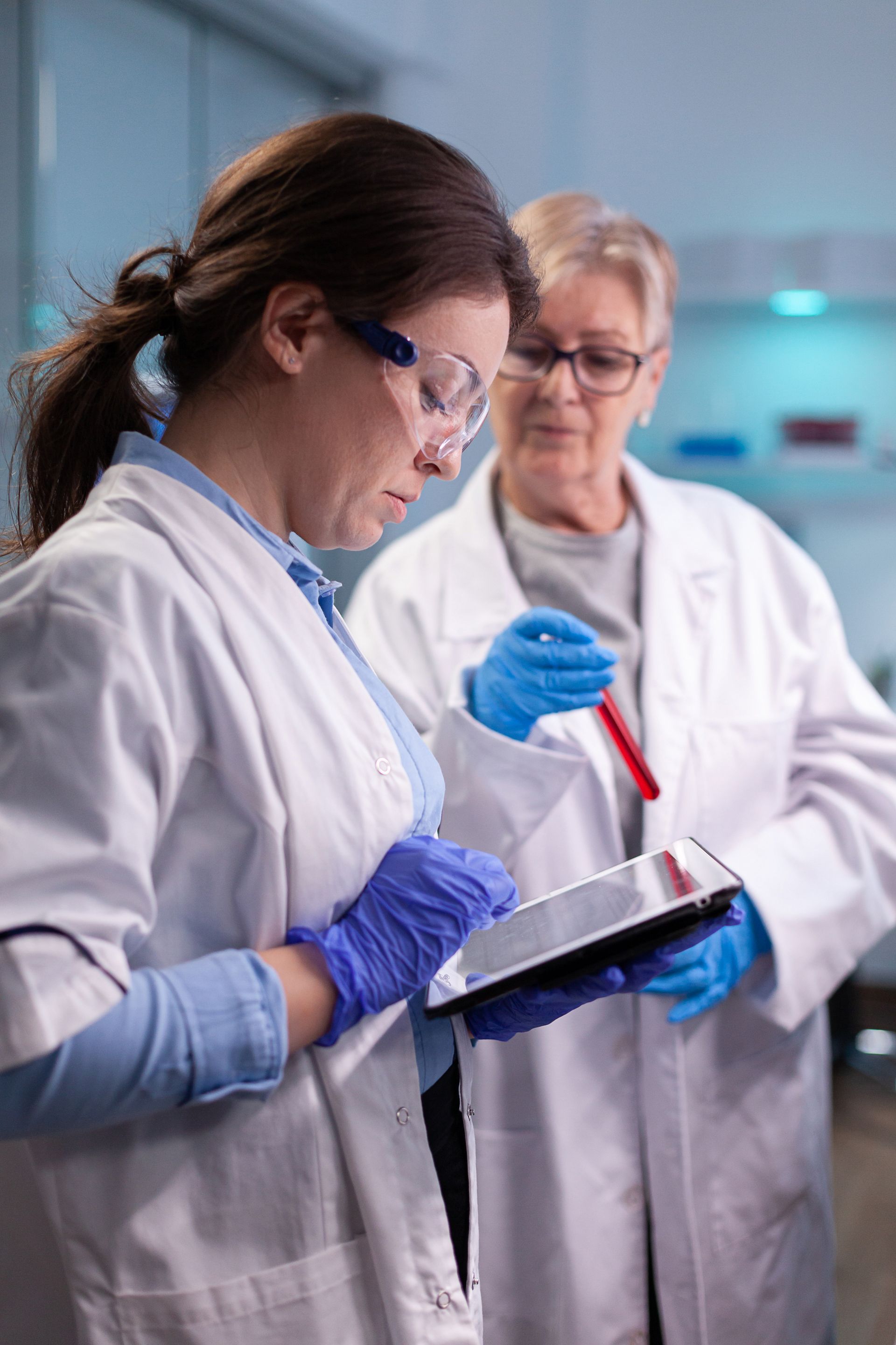 Two laboratory workers in white coats and blue gloves analyze data on a tablet while holding a test tube in a lab setting.
