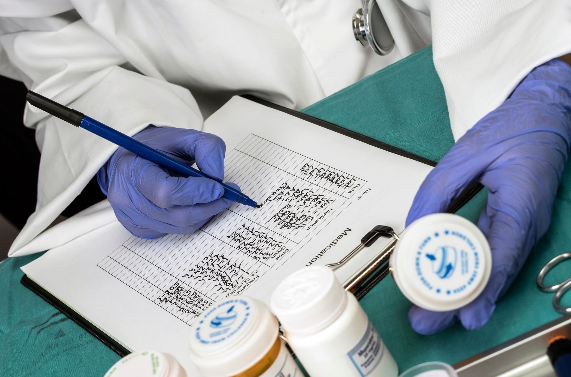 A gloved healthcare worker writes on a chart while holding a small medication container at a sterile station.