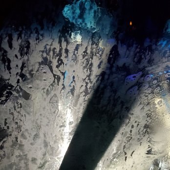 Inside a car wash, soapy foam coats a window, lit by blue and orange lights, with a wiper blade visible.