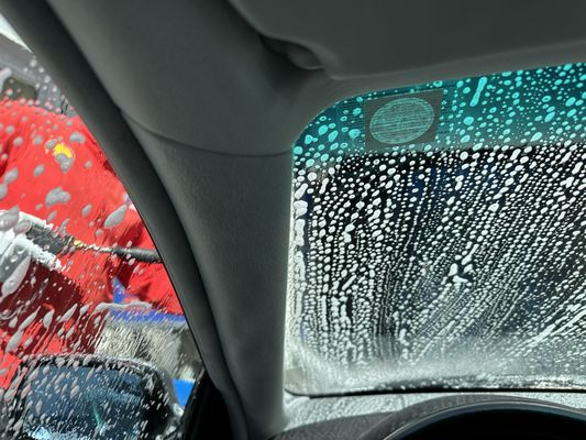 Car windshield covered in soapy water at a car wash; a person in a red jacket washes the vehicle.