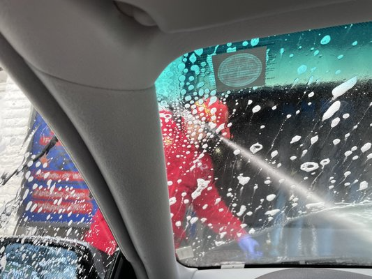 Inside a car at a car wash: person in red suit spraying soapy water onto the windshield.
