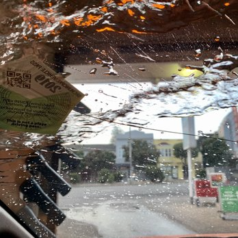View from inside a car wash, with water droplets on the windshield and buildings in the background. A sticker is visible.
