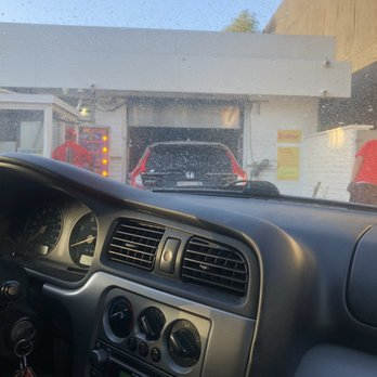 Interior car view at a car wash; red car inside, worker in red shirt, white building.