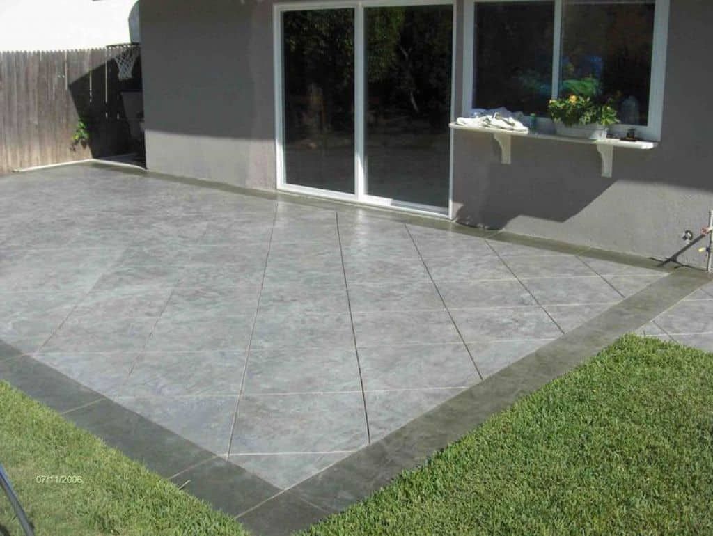 Gray concrete patio with border next to green grass and a house.