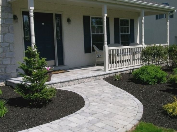 Curving paved walkway leading to a house with a porch and dark blue door, surrounded by dark mulch and greenery.