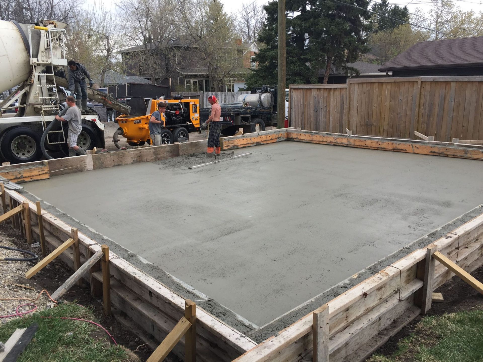 Concrete slab being poured, workers leveling concrete. Trucks and wooden forms visible.