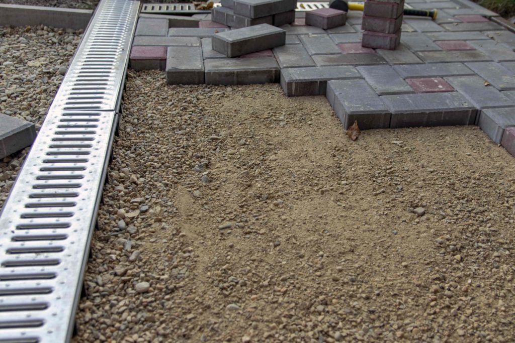 Construction of a paved area: bricks being laid on a bed of sand and gravel, with a drainage channel.
